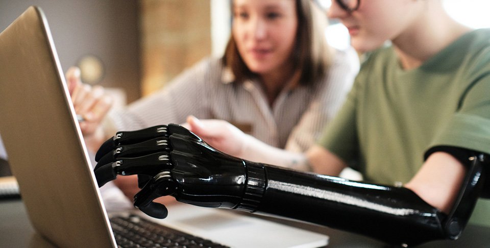 Close-up of woman with prosthesis arm pointing at monitor of laptop and discussing with her colleague at office desk